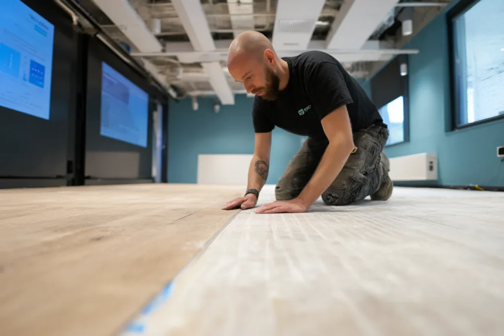 Monteur van E=MC² Facilitair die een nieuwe houten vloer nauwkeurig aanbrengt tijdens een vloerrenovatie in een modern kantoor.